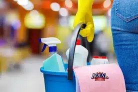 yellow-gloved hand carrying a blue bucket containing cleaning solutions and napkins